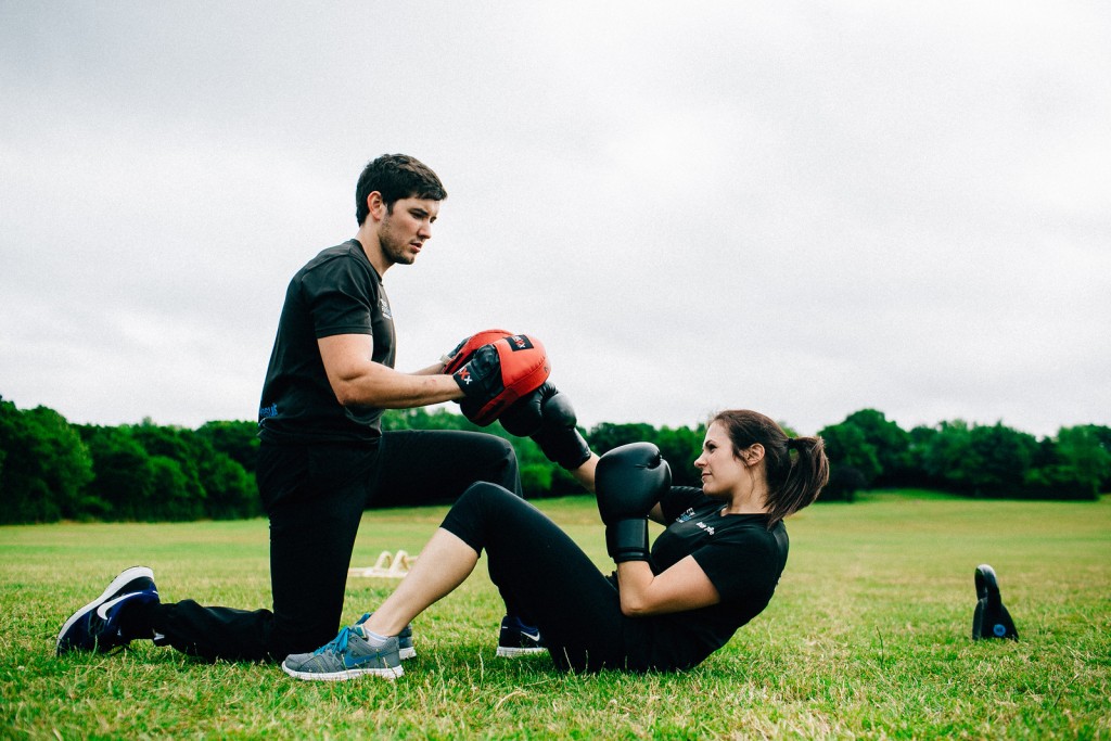 A close-up shot of a client smiling while wearing boxing gloves and throwing a punch toward focus pads. The image captures the energy, motivation, and enjoyment of a dynamic outdoor personal training session.